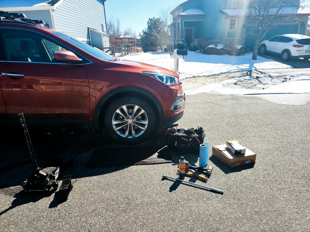 Denver mobile mechanic service performing car repair on a red Hyundai Santa Fe in a residential neighborhood with snow on the ground. Tools and parts are visible.