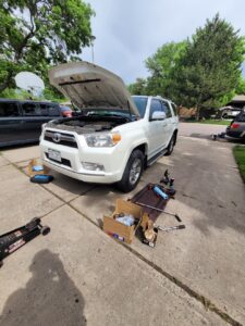 Mobile mechanic tools and equipment set up on a Denver driveway for on-site car repair, with a white SUV’s hood open and a jack, wrenches, and parts nearby.
