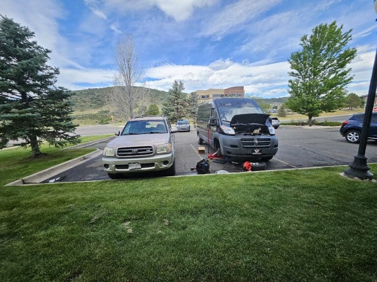 Automobile repair being performed outdoors on a black van with its hood open at a parking lot, featuring a silver car nearby and a scenic background of trees and hills.