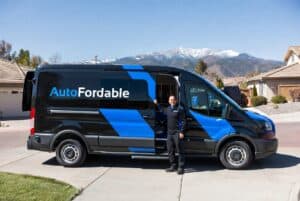 Mobile mechanic from AutoFordable standing next to a service van in a suburban neighborhood with snow-capped mountains in the background.