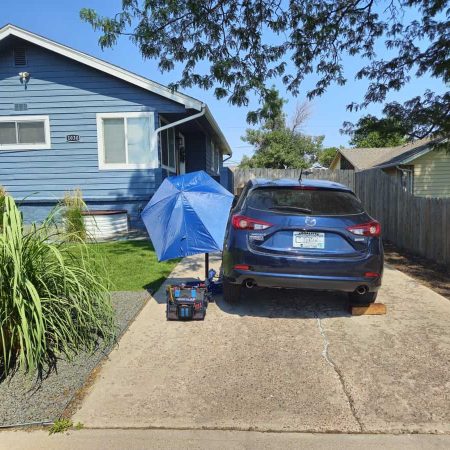 Car on driveway with tools and umbrella for mobile mechanic service in front of house.
