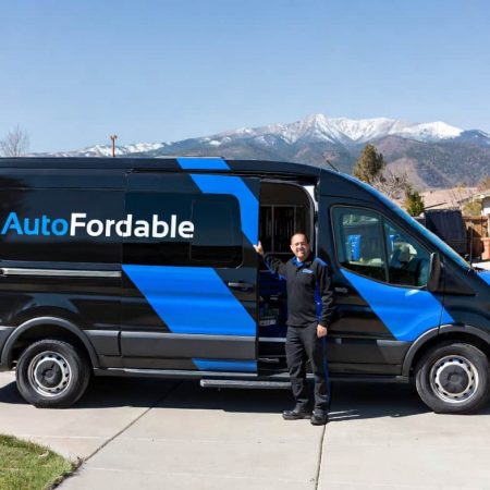 Mobile mechanic from AutoFordable standing next to a service van in a suburban neighborhood with snow-capped mountains in the background.