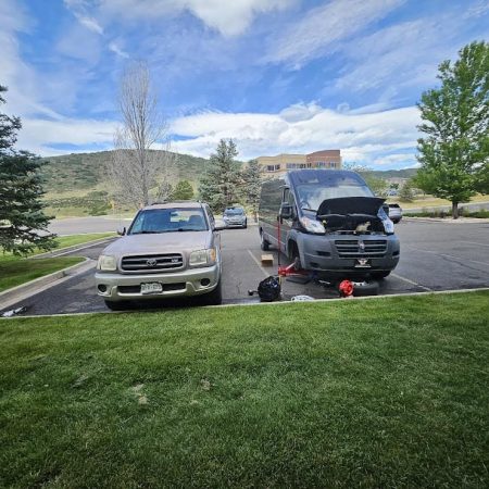 Automobile repair being performed outdoors on a black van with its hood open at a parking lot, featuring a silver car nearby and a scenic background of trees and hills.